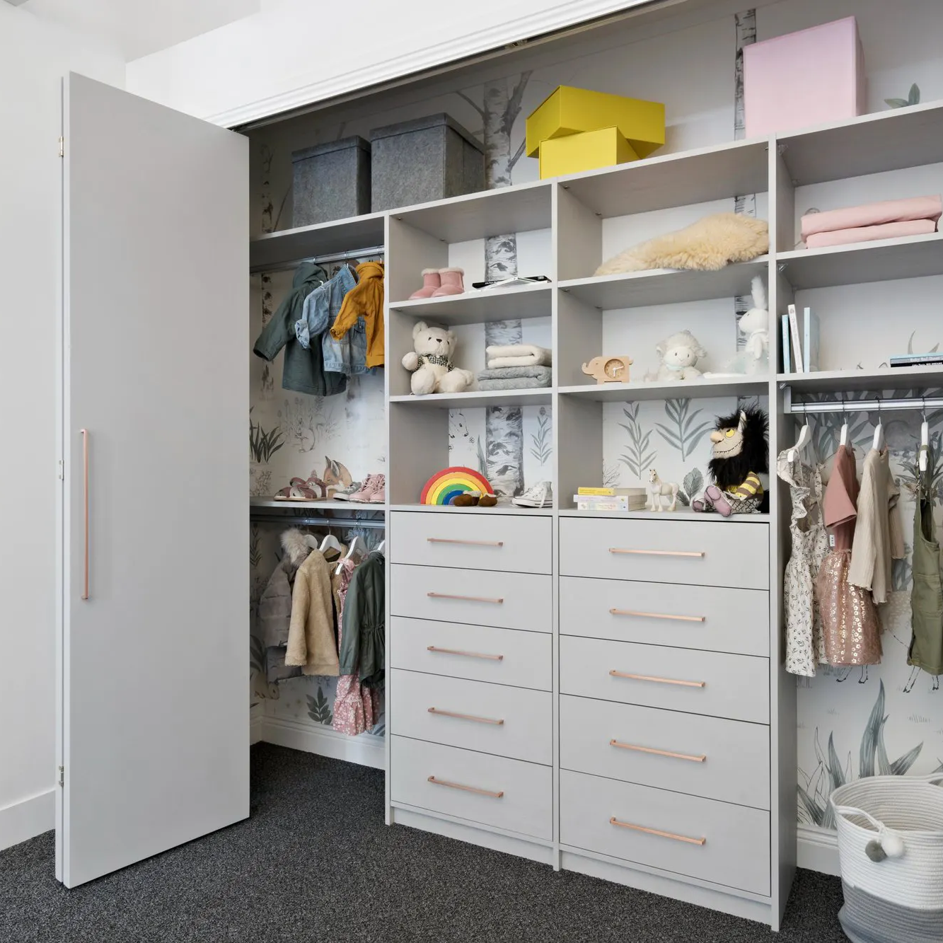 Image of a wall wardrobe or reach-in wardrobe with grey shelving and whimsical wallpaper. There are kids clothes and toys inside